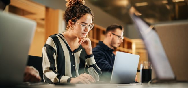 Portrait of Enthusiastic Hispanic Young Woman Working on Computer in a Modern Bright Office. Confident Human Resources Agent Smiling Happily While Collaborating Online with Colleagues.