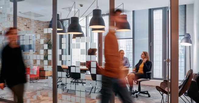 In the dynamic hustle and bustle of a business environment, a group of young business professionals walk down the corridor next to their office, while their colleagues collaborate inside, symbolizing teamwork and productivity.