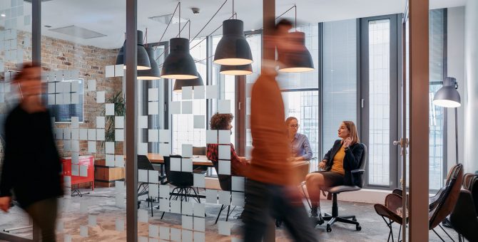 In the dynamic hustle and bustle of a business environment, a group of young business professionals walk down the corridor next to their office, while their colleagues collaborate inside, symbolizing teamwork and productivity.
